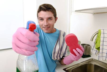 young attractive and happy man holding washing detergent spray bottle and cloth wearing rubber gloves smiling confident as aiming with hand gun playful enjoying housework and domestic dutyの写真素材