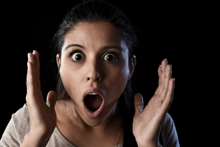 young beautiful scared Spanish woman in shock and surprise face expression astonished and amazed isolated on black background in primal facial emotion conceptの写真素材