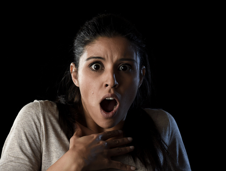 young beautiful scared Spanish woman in shock and surprise face expression astonished and amazed isolated on black background in primal facial emotion conceptの写真素材