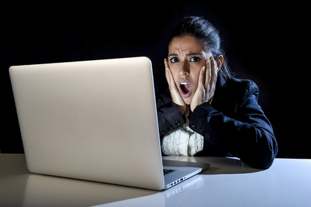 young hispanic business woman or student girl working in darkness on laptop computer late at night surprised in shock and angry in stress in crisis and failure concept isolated on black backgroundの写真素材