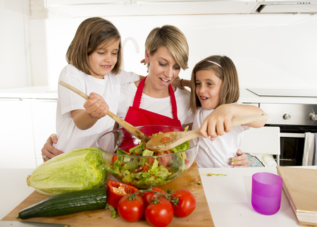 young mother in cook apron and sweet beautiful twin daughters cooking preparing together salad playful at home kitchen in healthy vegetable nutrition and homecook conceptの写真素材