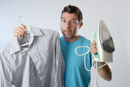 young attractive and frustrated man holding iron and shirt stressed and tired in bored and lazy face expression in male ironing going wrong and domestic work concept isolated even backgroundの写真素材