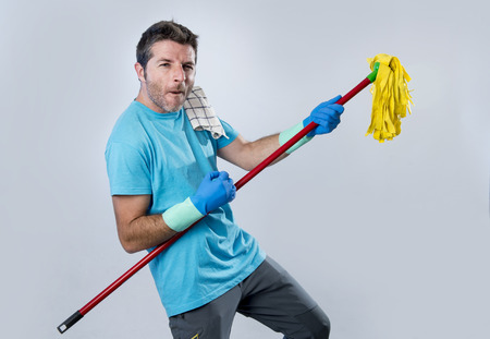 young domestic service man or happy husband cleaning home playing with mop a song on air guitar having fun enjoying isolated on even background in housekeeping and home cleaning conceptの写真素材