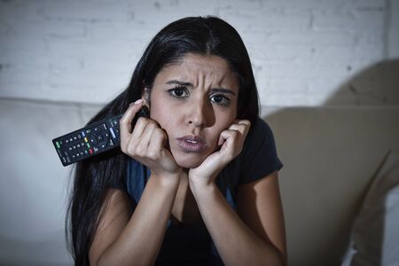Spanish woman at home sofa couch in living room watching television very concentrated and focused paying attention to tv news or program in disbelief face expression shocked and amazedの写真素材