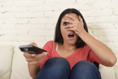young beautiful latin woman sitting at home sofa couch in living room watching television scary horror movie or horrible news scared and excited covering her eyes holding remote controlの写真素材
