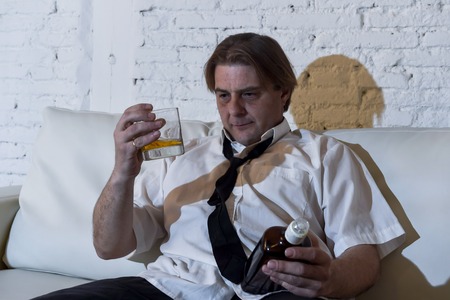 sad depressed alcoholic businessman on his 40s with loose necktie looking wasted and drunk drinking whiskey and smoking at home living room couch in alcoholism problem and alcohol addictionの写真素材
