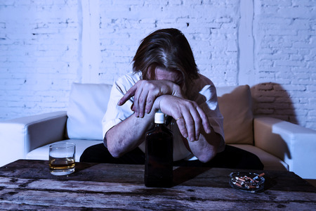 sad depressed alcoholic businessman on his 40s with loose necktie looking wasted and drunk drinking whiskey and smoking at home living room couch in alcoholism problem and alcohol addictionの写真素材
