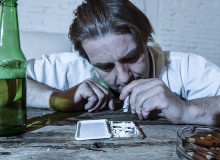 wasted alcoholic and drug addict man with loose tie snorting cocaine and drinking beer bottles at home living room couch in toxic substance addiction and abuse conceptの写真素材