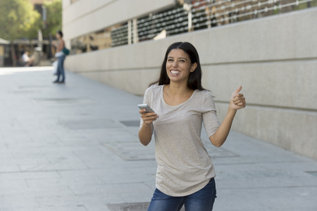 young beautiful and happy latin woman on urban street city background using internet on mobile phone texting and browsing social media in online communication concept giving thumb upの写真素材