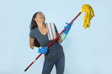 young attractive happy Latin woman in washing gloves holding mop having fun singing and playing air guitar excited and cheerful isolated on blue background in house cleaning and domestic work conceptの写真素材