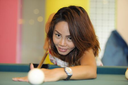 young pretty and happy Asian girl playing snooker holding stick at pool table in night club or bar...
