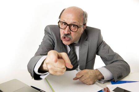 60s bald senior office boss man furious and angry gesturing upset and mad sitting on desk with paperwork in business and job problems and stress concept isolated on white backgroundの写真素材