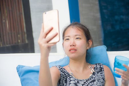 young sweet and pretty Asian Chinese woman sitting at beach holiday resort taking selfie picture on mobile phoneの写真素材