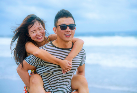 young beautiful Asian Chinese couple with boyfriend carrying woman on her back and shoulders at the beach smiling happy in love enjoying holidays and joyful in romantic trip and romance conceptの写真素材