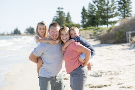 young happy beautiful Caucasian family having holidays on the beach smiling with mother and father carrying little son and young adorable daughter on their back in vacation lifestyle and love conceptの写真素材