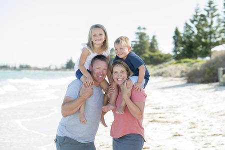 young happy beautiful Caucasian family having holidays on the beach smiling with mother and father carrying little son and young adorable daughter on shoulders in vacation lifestyle and love conceptの写真素材
