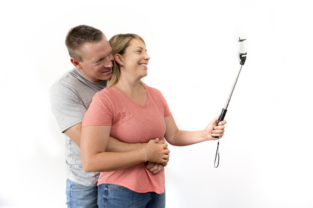 young beautiful happy and attractive romantic couple with husband and wife or girlfriend and boyfriend taking selfie self portrait photo with stick and mobile phone camera isolated on white backgroundの写真素材