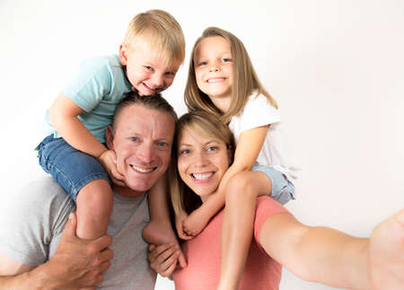 lovely young couple taking selfie photo self portrait with stick and mobile phone carrying son and daughter on shoulders posing happy smiling isolated on white in family lifestyle conceptの写真素材