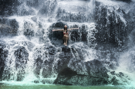 young beautiful and sweet Asian woman in bikini getting body wet under stream of natural amazing waterfall sitting on rock feeling purity and freedom in secret nature beauty travel destinationの写真素材