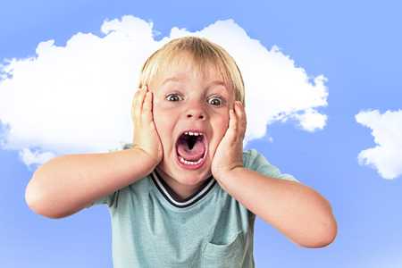 portrait of 3 years old little blond kid with mouth open screaming surprised or scared holding his head with his small hands isolated on blue sky background in facial expression and emotions conceptの写真素材