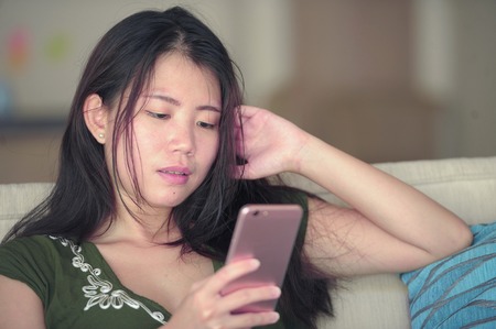 young beautiful and relaxed Asian Chinese woman lying at home living room sofa couch using internet...