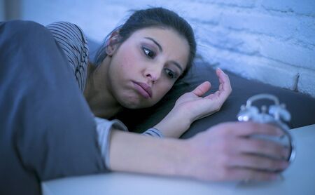 young beautiful and annoyed latin woman waking up upset to the sound of alarm clock early  morning feeling lazy to get up or suffering insomnia and sleeping disorderの写真素材