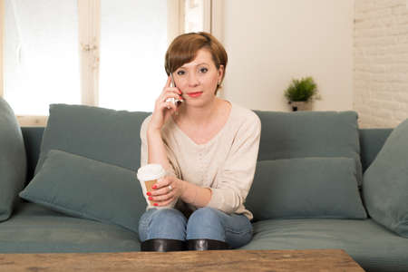 young attractive and happy red hair woman sitting at home sofa couch drinking coffee talking on...
