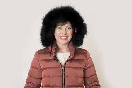 young attractive and happy red hair Caucasian woman on her 20s or 30s posing cheerful and smiling wearing warm winter jacket and fur hood isolated on grey background in seasonal fashion の写真素材