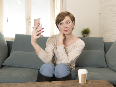 young attractive and happy red hair woman sitting at home sofa couch drinking coffee taking selfie picture with mobile phone camera in internet social media addiction and lifestyle conceptの写真素材