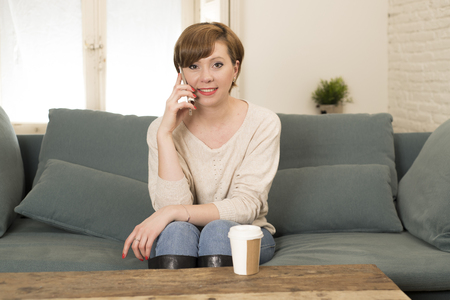 young attractive and happy red hair woman sitting at home sofa couch drinking coffee talking on mobile phone relaxed in communication and lifestyle conceptの写真素材