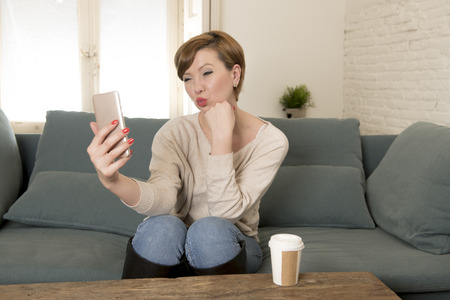 young attractive and happy red hair woman sitting at home sofa couch drinking coffee taking selfie picture with mobile phone camera in internet social media addiction and lifestyle conceptの写真素材