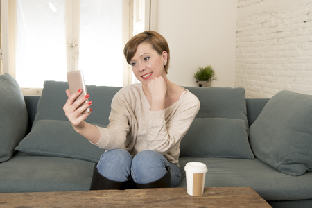 young attractive and happy red hair woman sitting at home sofa couch drinking coffee taking selfie picture with mobile phone camera in internet social media addiction and lifestyle conceptの写真素材