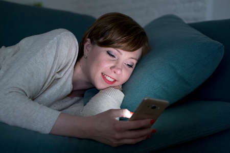 young pretty and happy red hair woman on her 20s or 30s lying on home couch or bed using mobile phone late at night smiling relaxed in internet addiction and social media obsession conceptの写真素材