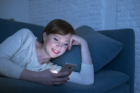young pretty and happy red hair woman on her 20s or 30s lying on home couch or bed using mobile phone late at night smiling relaxed in internet addiction and social media obsession conceptの写真素材