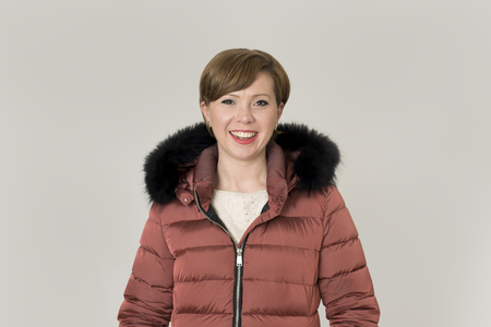 young attractive and happy red hair Caucasian woman on her 20s or 30s posing cheerful and smiling wearing warm winter jacket with fur hood isolated on grey background in seasonal fashion conceptの写真素材