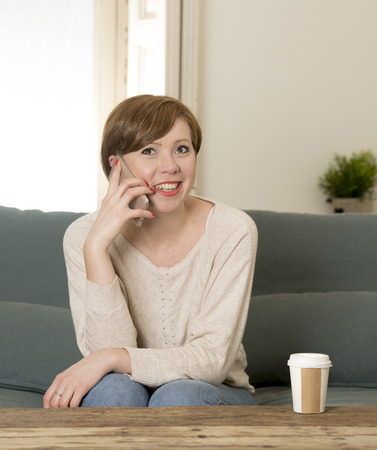 young attractive and happy red hair woman sitting at home sofa couch drinking coffee talking on mobile phone relaxed in communication and lifestyle conceptの写真素材