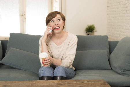 young attractive and happy red hair woman sitting at home sofa couch drinking coffee talking on mobile phone relaxed in communication and lifestyle conceptの写真素材