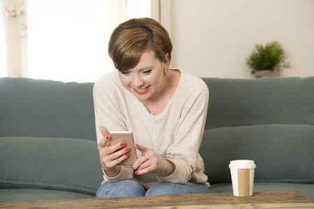 young attractive and happy red hair woman sitting at home sofa couch drinking coffee using internet dating app on mobile phone relaxed in communication and lifestyle conceptの写真素材