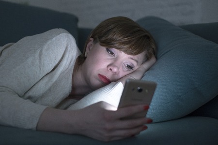 portrait of young woman 30s lying on bed couch late at night at home using social media app on mobile phone tired and sleepy in internet addiction conceptの写真素材