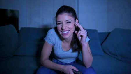young beautiful and happy hispanic woman on her 30s holding TV remote enjoying at home living room couch watching television show comedy movie smiling having fun relaxed in lifestyleの写真素材