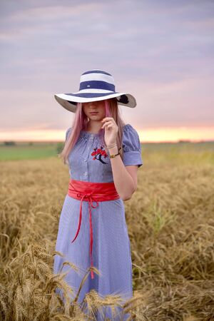 A girl in a blue dress and a light hat stands in the middle of a wheat field yellowの写真素材