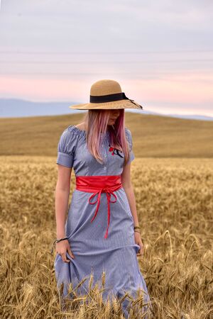 A girl in a blue dress and a light hat stands in the middle of a wheat field yellowの写真素材