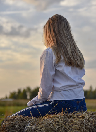 Girl with beautiful long hair at sunset sitting on a haystack back to the photographer dreamilyの写真素材