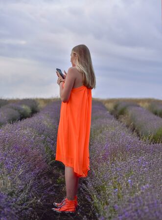 A girl in a bright orange dress and standing in a field of lavender at dawn holding a smartphone skyの写真素材