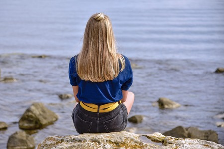 A girl in a blue jacket with her hair down sits on a stone by the calm sea and looks into the distance with her back to.の写真素材