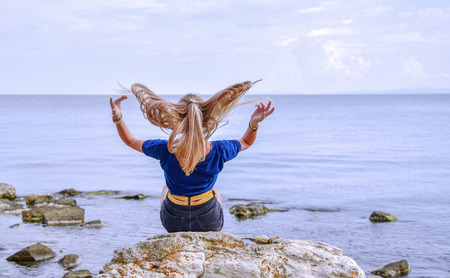 A girl in a blue jacket with her hair down sits on a stone by the calm sea and looks into the distance with her back to.の写真素材
