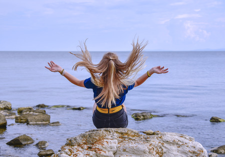 A girl in a blue jacket with her hair down sits on a stone by the calm sea and looks into the distance with her back to.の写真素材