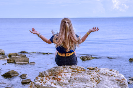 A girl in a blue jacket with her hair down sits on a stone by the calm sea and looks into the distance with her back to.の写真素材