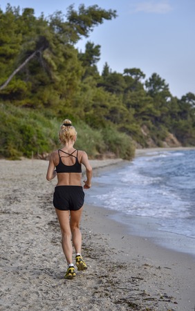 Sporty girl in black swimsuit runs along the sea in summer morning jog back to the cameraの写真素材