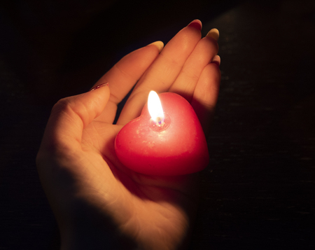 Women's hand holding a burning red candle in the dark lightの写真素材